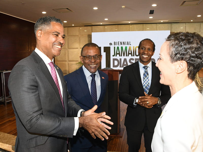 Four professionals in suits smile and greet each other in a conference room, with an '11th Biennial Jamaica Diaspora' banner behind them.