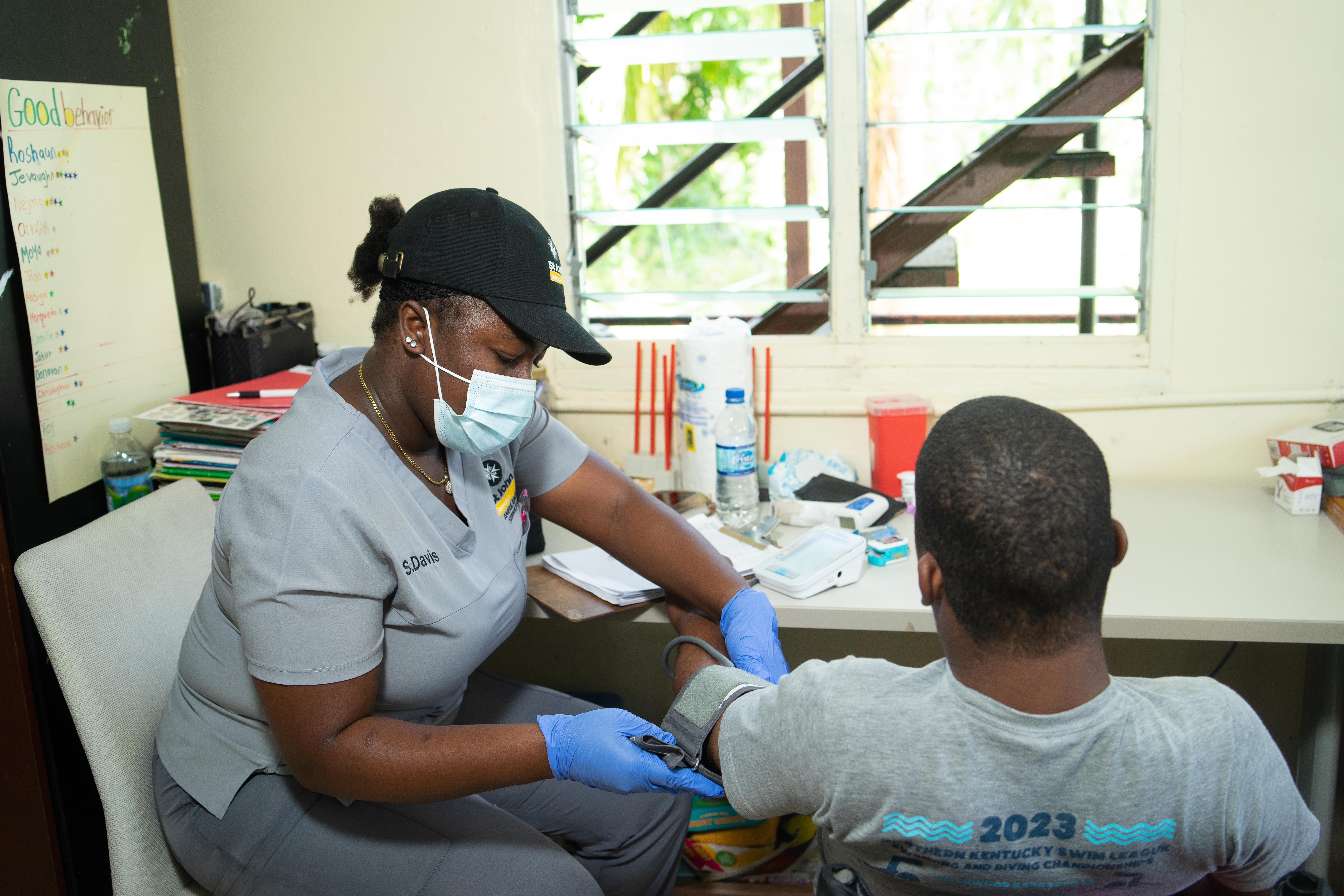 Healthcare worker in scrubs and mask draws blood from a seated patient in a clinic, with supplies on the desk nearby.