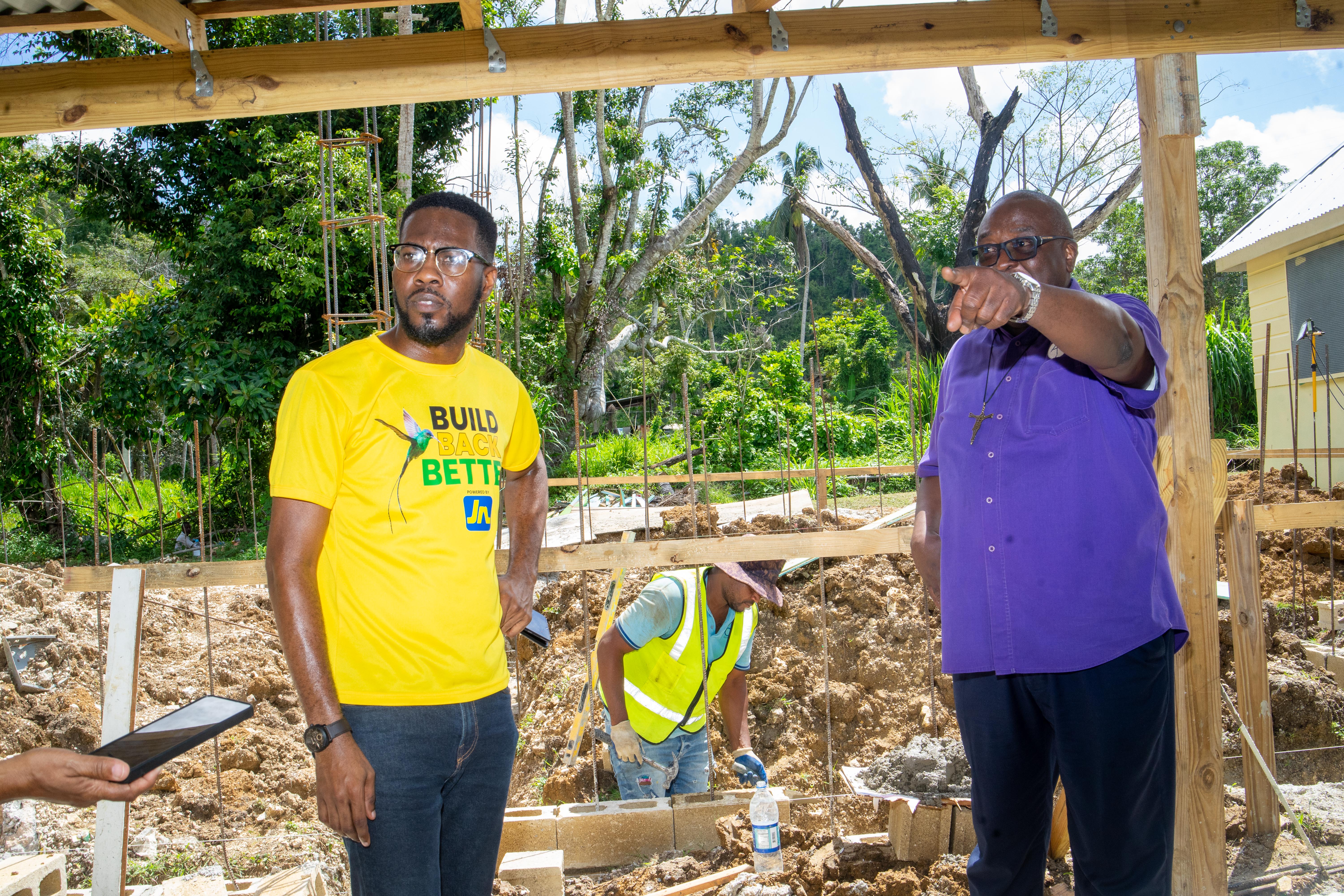 Two men on a construction site: a man in a yellow shirt reading 'BUILD BACK BETTER' and a man in a purple shirt pointing, with wooden framing and workers in the background.