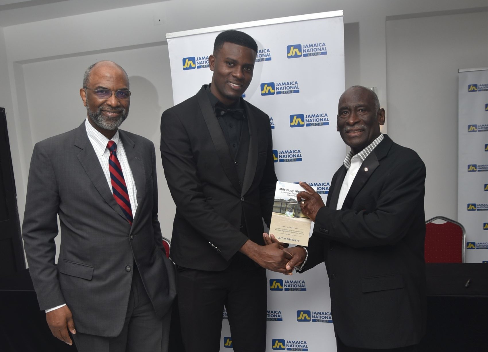 Three men in suits at a Jamaica National event; the center man shakes hands with the man on the right while receiving a certificate.
