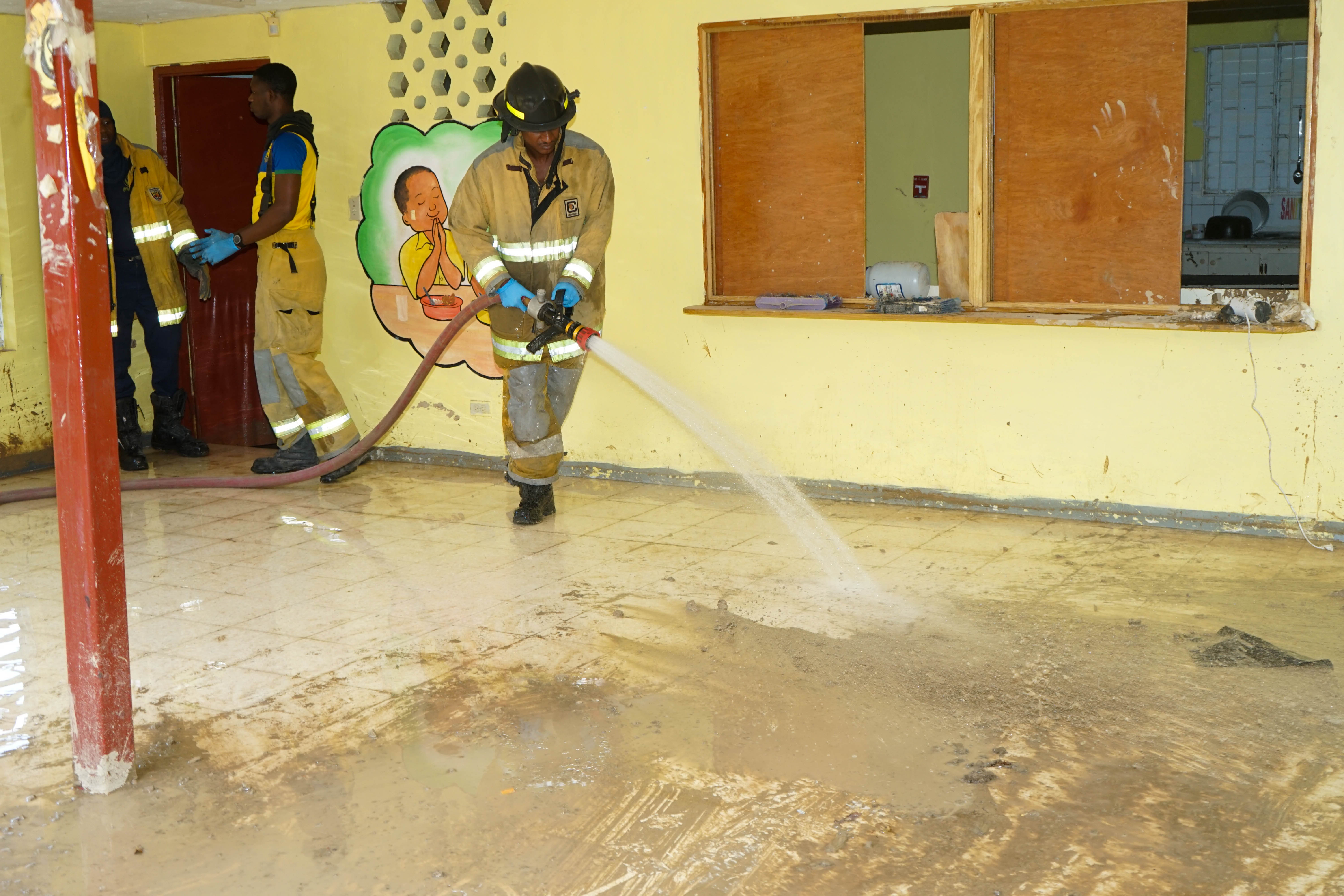 Firefighters in yellow turnout gear spray water inside a yellow-walled room, with a mural of a praying child on the wall behind them.