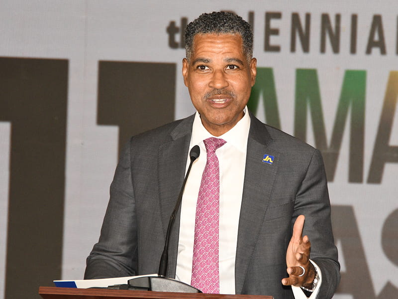 Man in a gray suit and pink tie delivers a speech at a podium with a microphone, gesturing with his right hand.