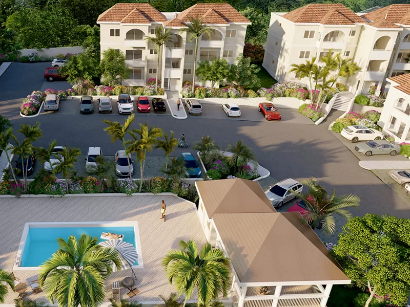 Aerial view of a tropical residential complex with beige buildings, red-tile roofs, a pool area with lounge chairs, palm trees, and a parking lot full of cars.
