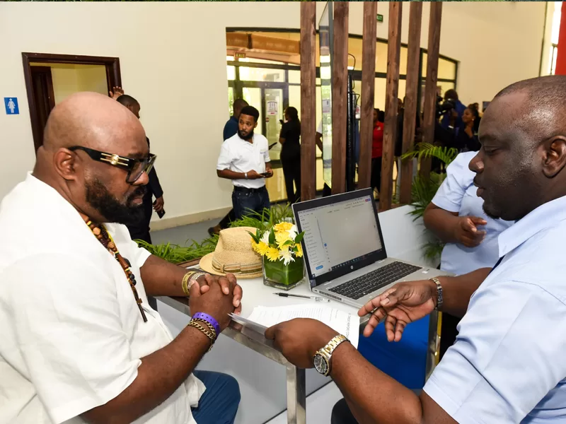 ParkAve1 - The Jamaica National Group Limited Two men at a reception desk reviewing paperwork; laptop open, vase of yellow flowers, and a straw hat on the counter, with people mingling in the background.