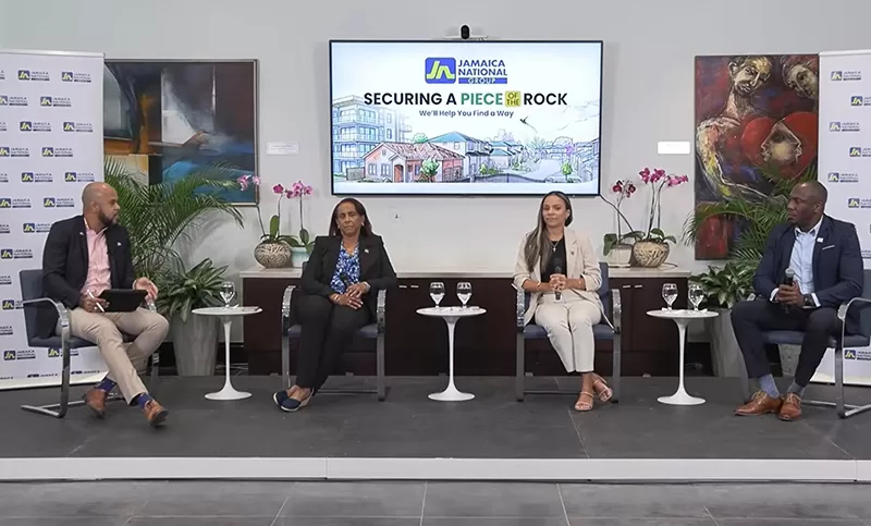 Panel discussion with four speakers seated on stage in a conference room, microphones in hand, and a screen behind them displaying Jamaica National branding.