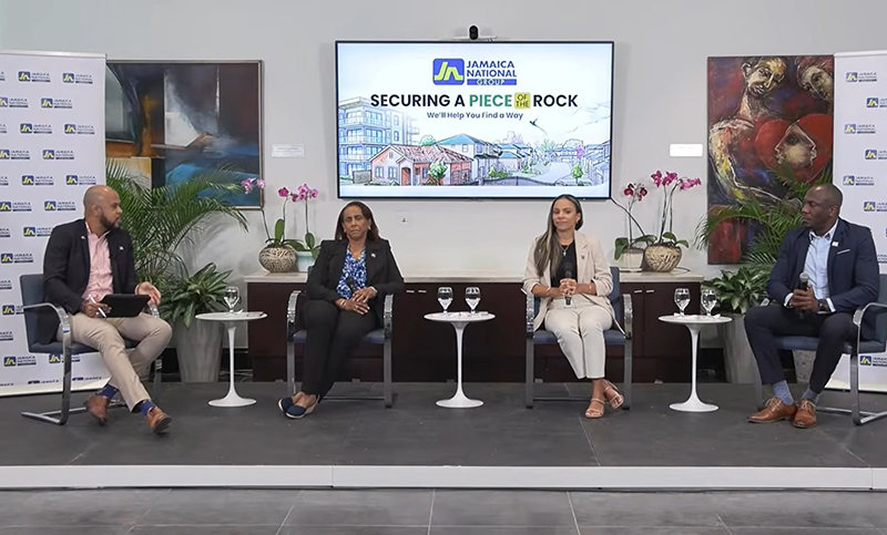 Panel discussion with four speakers seated on stage in a conference room, microphones in hand, and a screen behind them displaying Jamaica National branding.