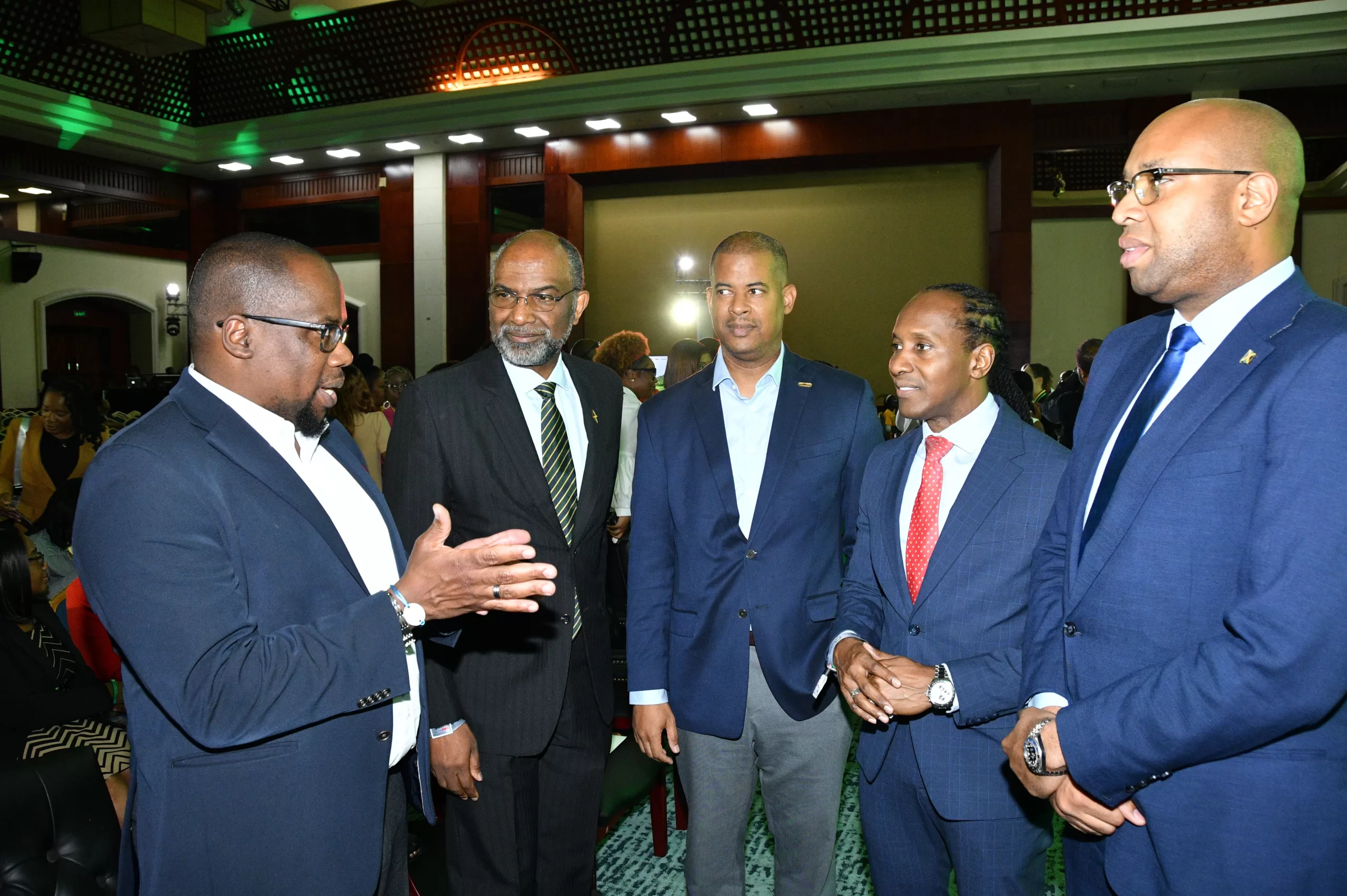Five men in dark blue suits chatting in a conference hall, one gesturing with his hands as they stand in a semi-circle.
