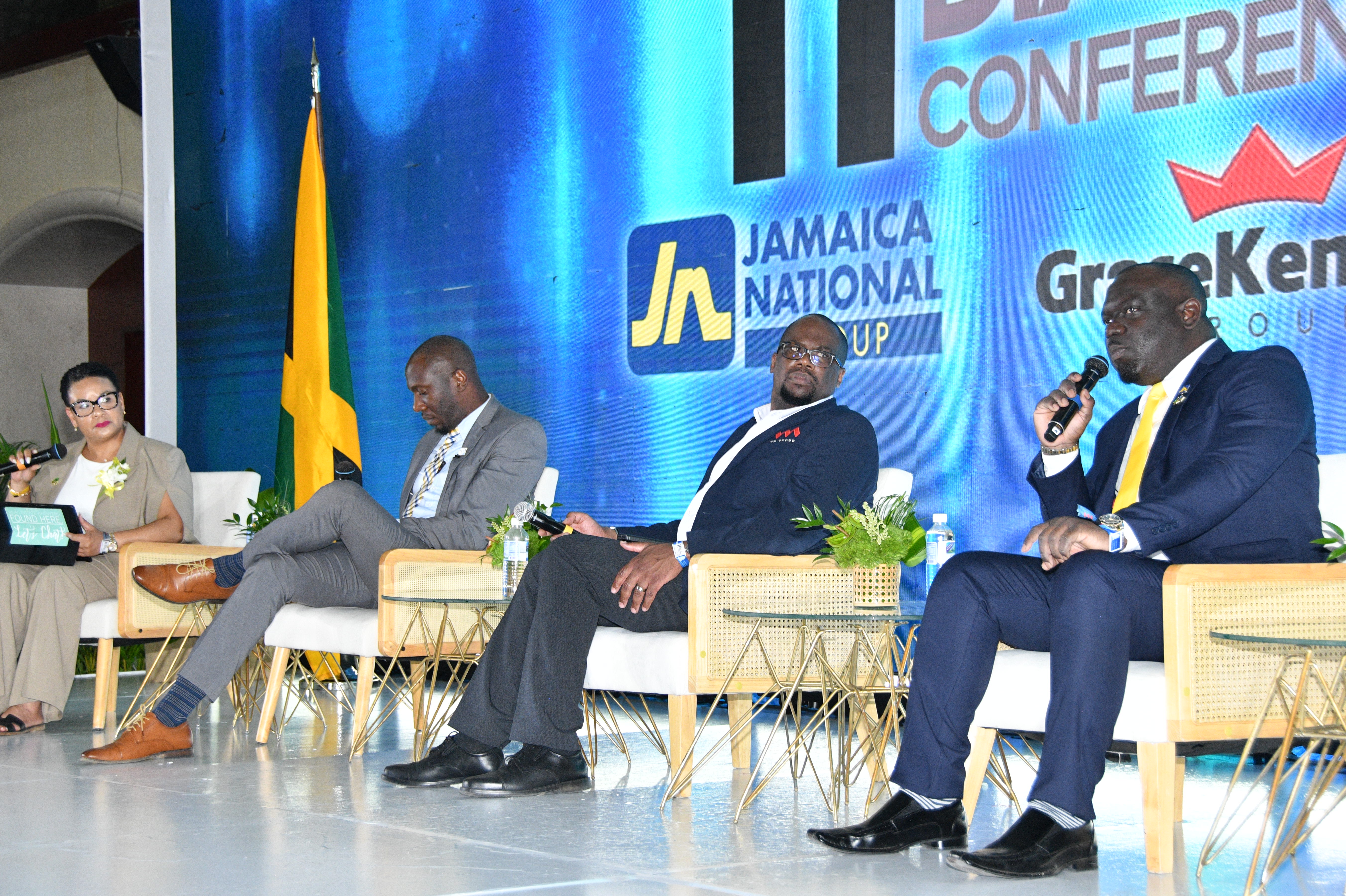 Panel discussion on a stage with four speakers in business attire; a Jamaican flag and a blue conference backdrop are visible behind them, microphones in hand.