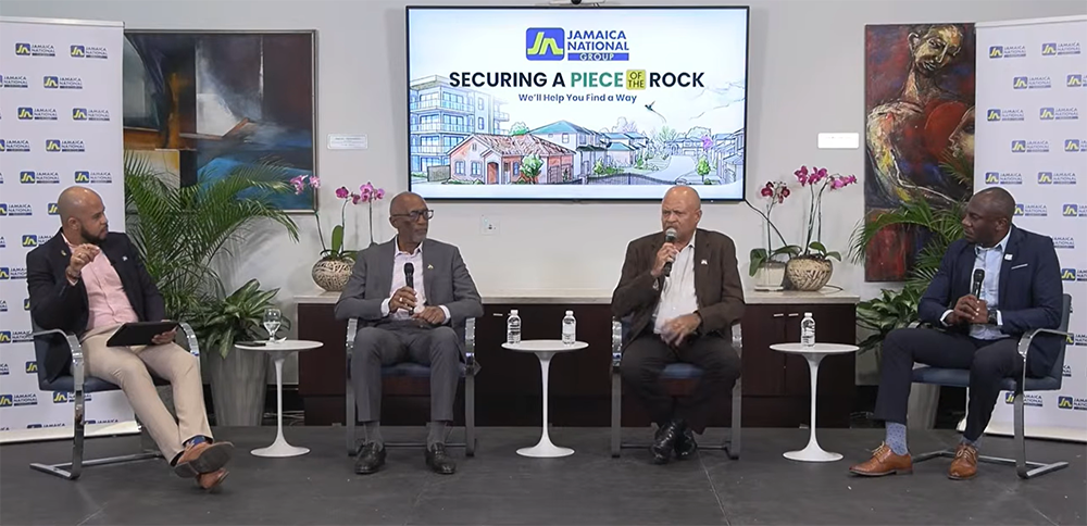 Panel discussion with four men in business attire seated on stage, microphones in hand; screen behind reads 'Securing a Piece of the Rock' with Jamaica National branding.