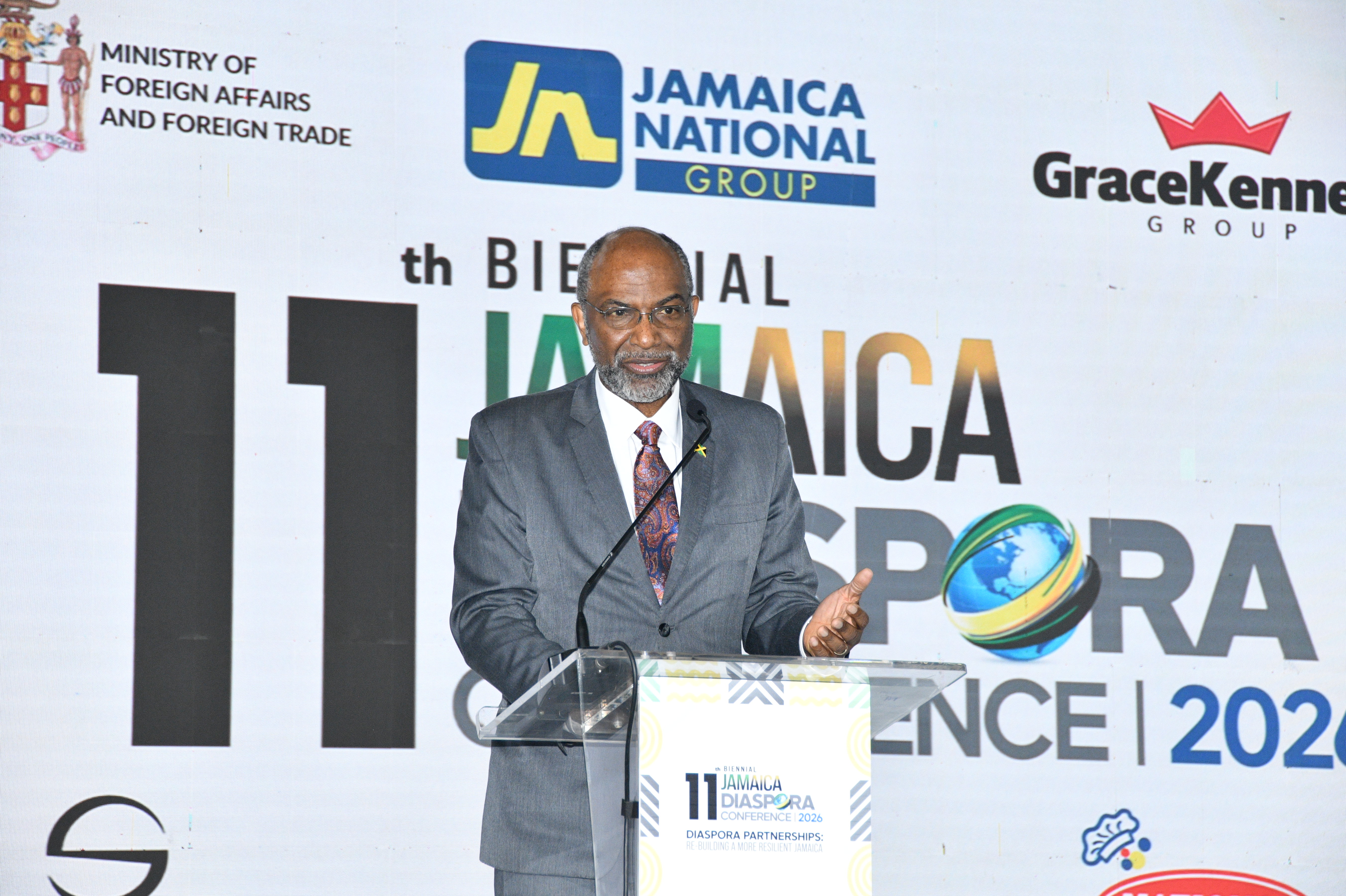 Speaker in a gray suit delivering a speech at a podium, with a Jamaica Diaspora Conference backdrop and partner logos in the background.