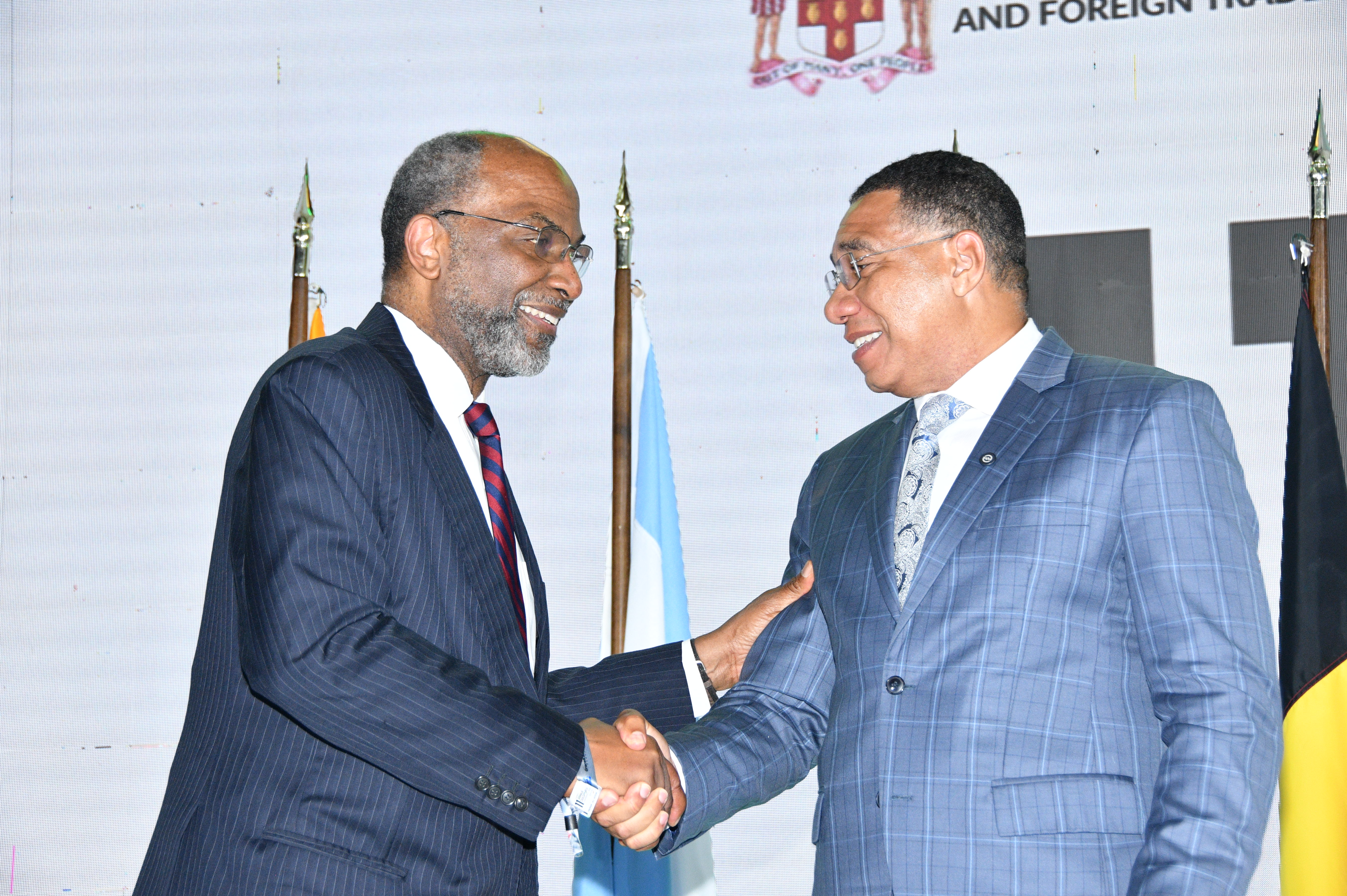 Two men in dark business suits shake hands and smile at a formal event, flags visible in the background.