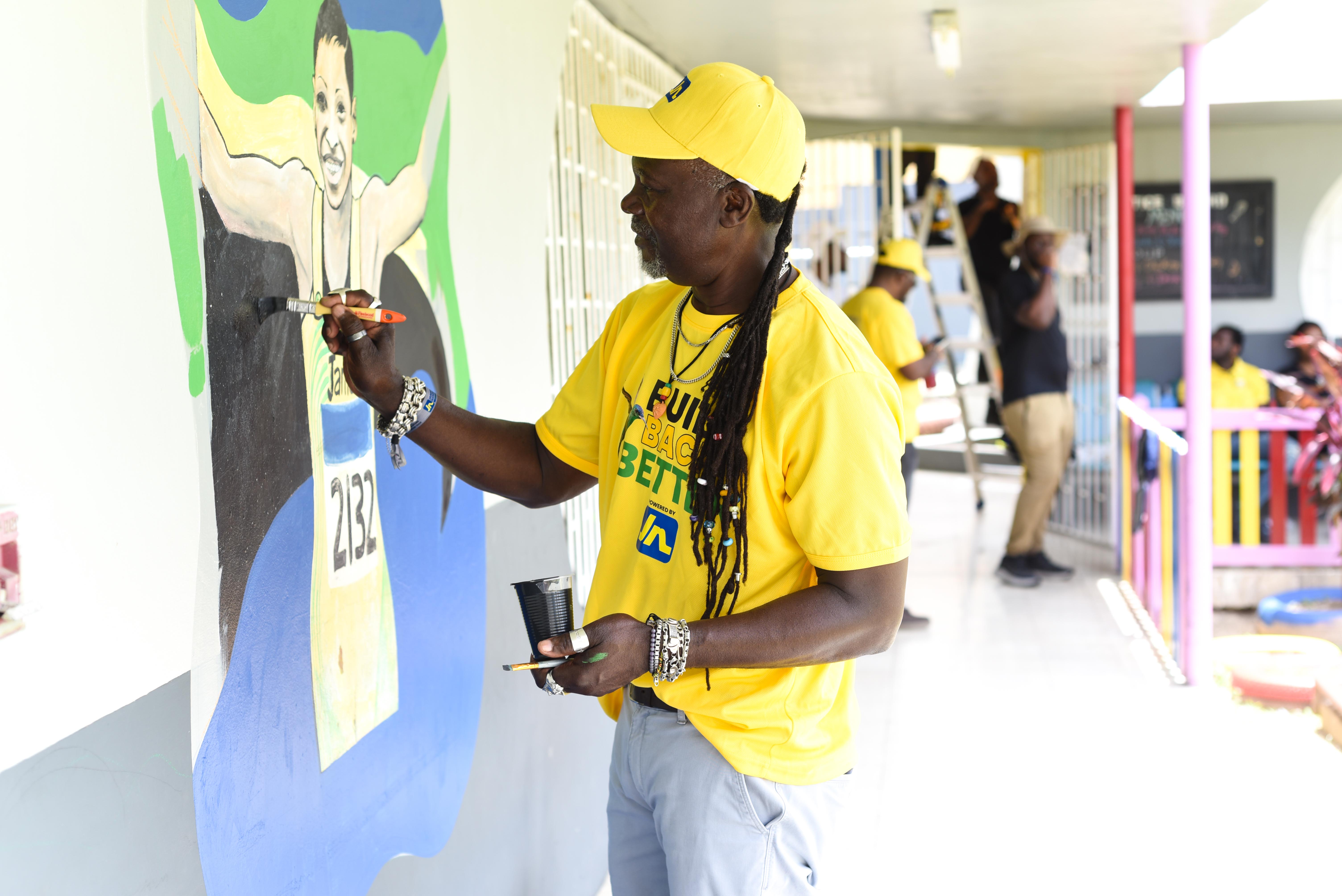 SNP_5342 - The Jamaica National Group Limited A man in a yellow cap and shirt paints a colorful mural on a wall outdoors during a community event.