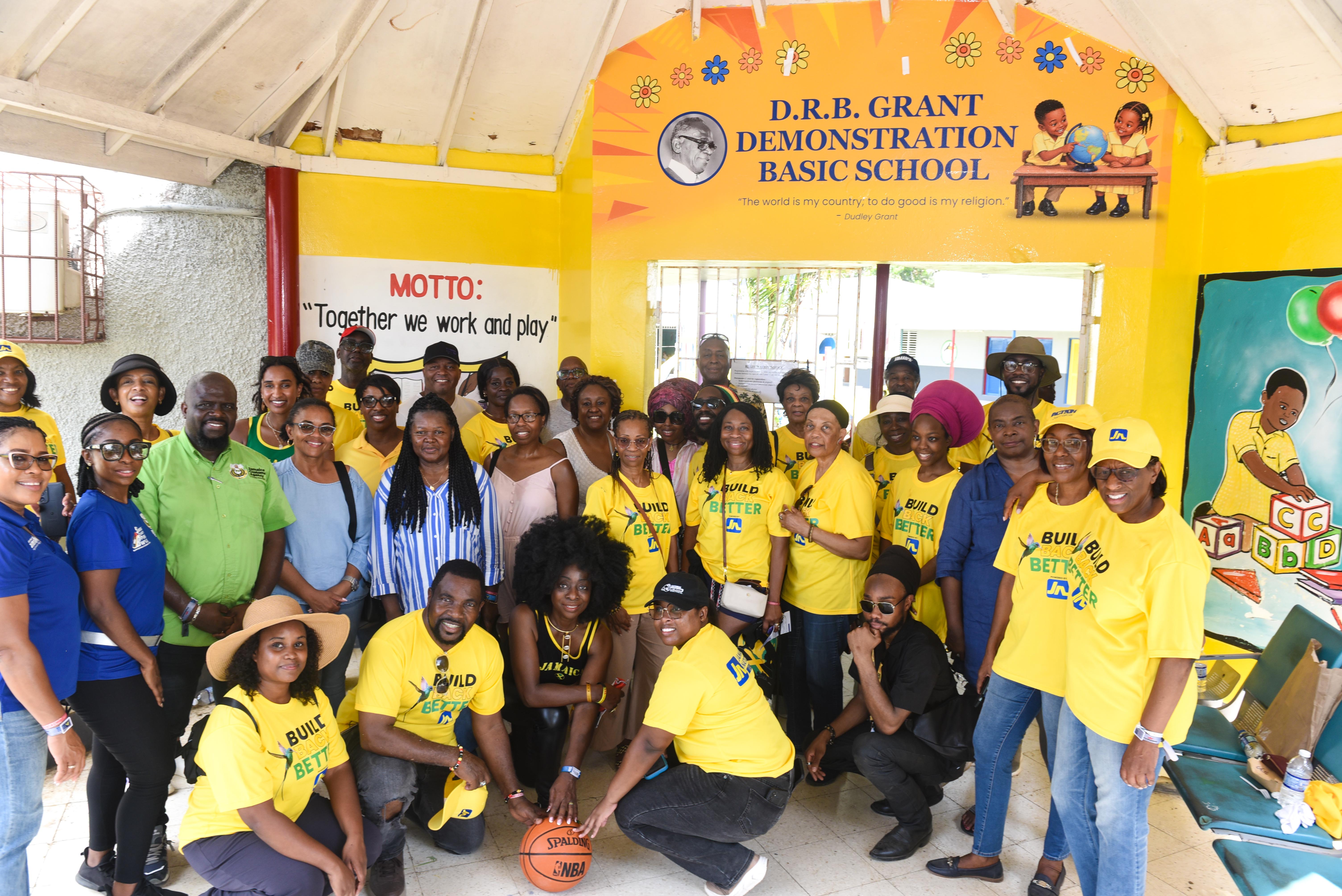 Group of smiling adults and a few children in yellow shirts posing for a group photo inside a colorful school lobby, some with hats and sunglasses.