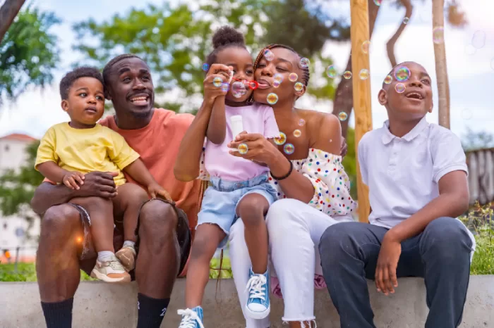 Family of five sitting on a low wall outdoors, smiling and blowing bubbles together on a sunny day.