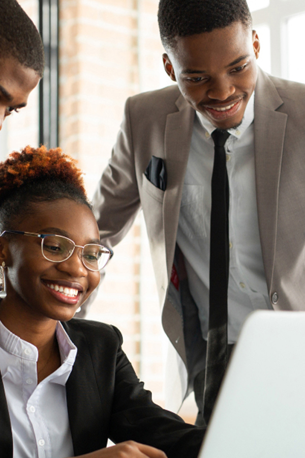 Three colleagues in business attire gathered around a laptop, smiling as they work together in a bright office.