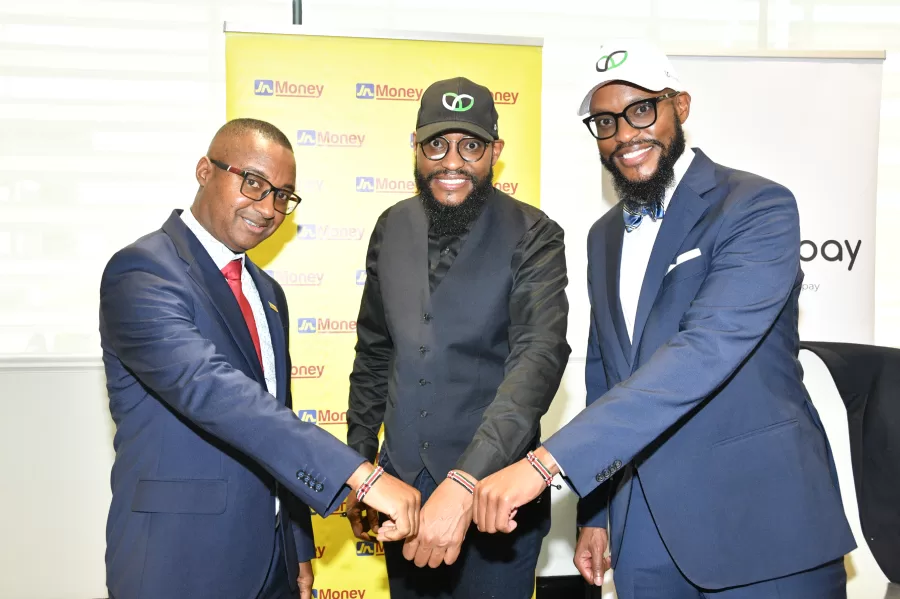 Three men in suits fist-bump together at a business event, with JN Money banners in the background.