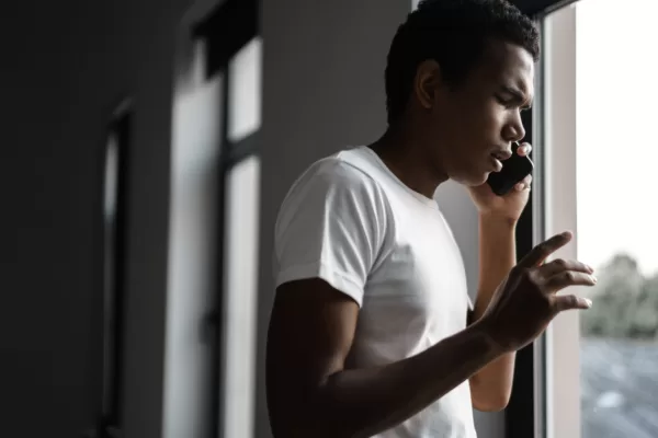 Man in a white t-shirt talking on a cellphone by a window, looking outward with a thoughtful expression in a dim hallway.