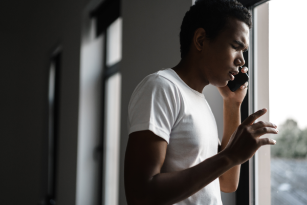 Man in a white t-shirt talking on a cellphone by a window, looking outward with a thoughtful expression in a dim hallway.
