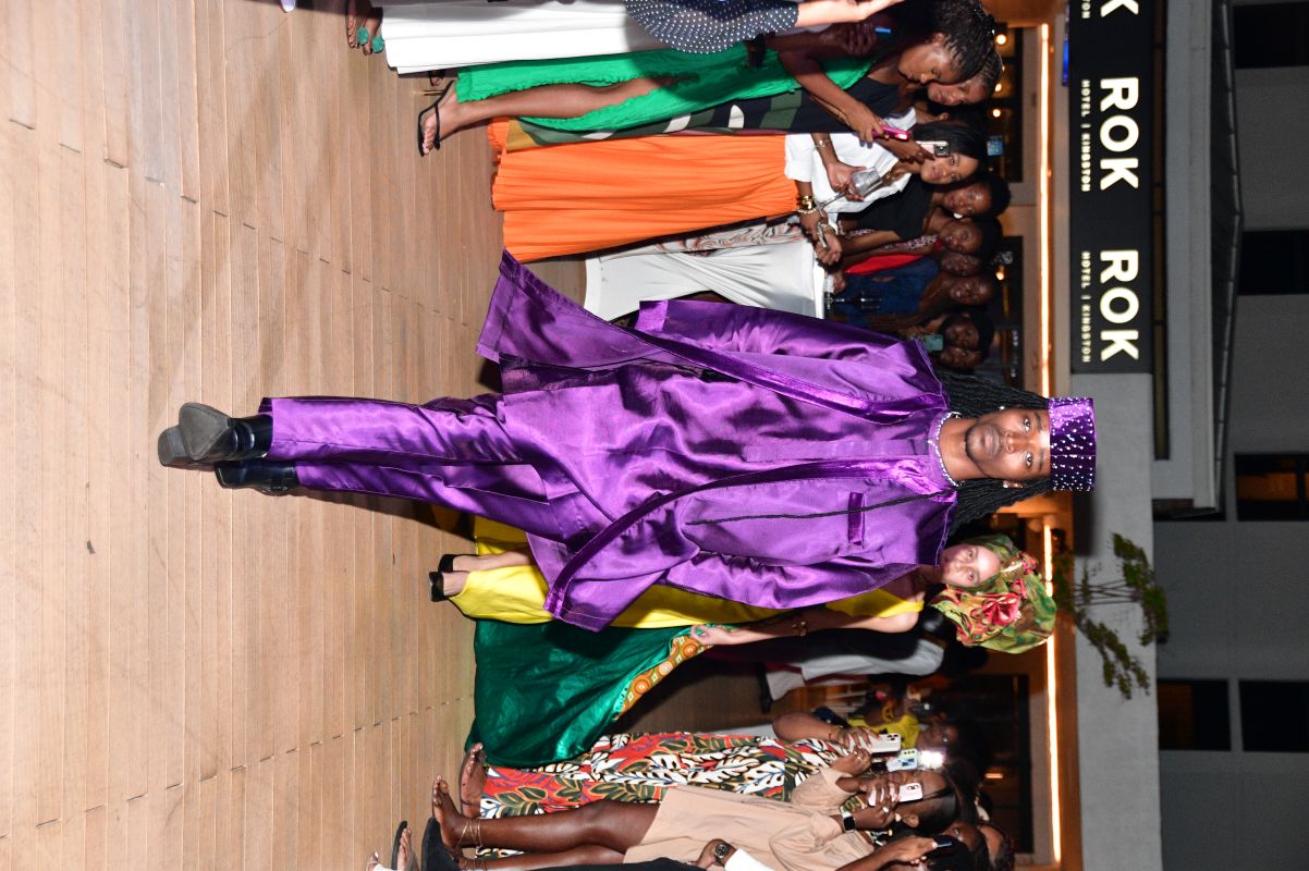 Male model in a metallic purple traditional outfit walking on a runway at night, surrounded by spectators.