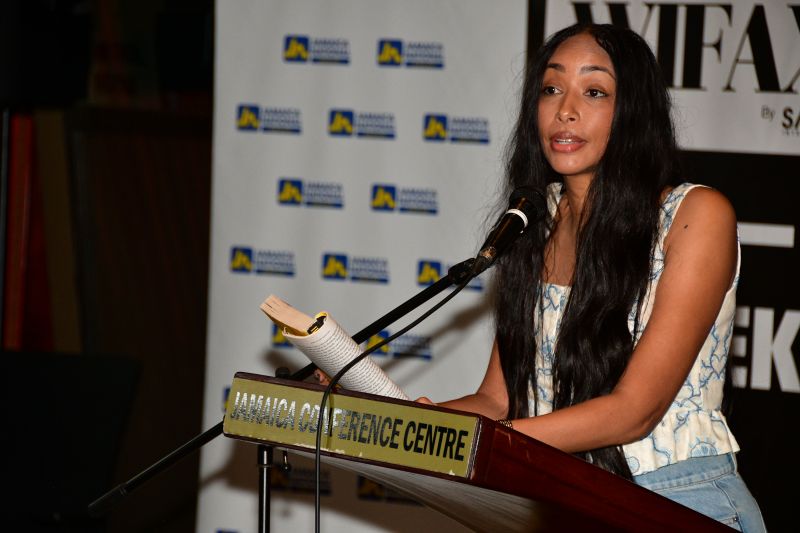 Woman with long dark hair speaks at a podium with a microphone at a Jamaica Conference Centre event, holding notes as she addresses the audience.