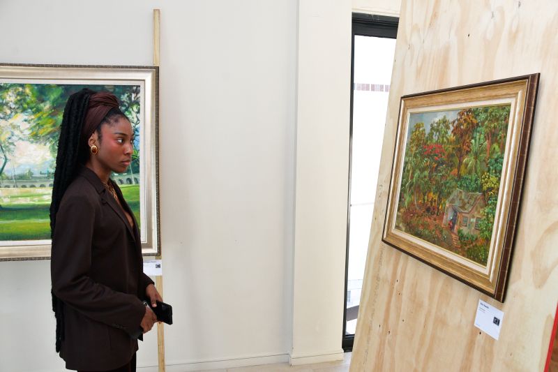 Woman with braids in a brown blazer viewing a framed landscape painting in an art gallery.