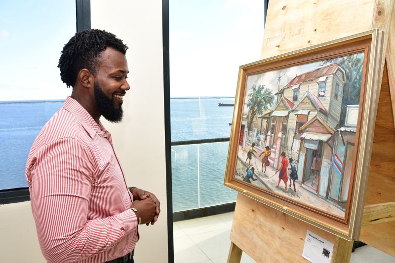 A man in a pink striped shirt smiles while looking at a framed painting on an easel near a large window overlooking water and a distant shoreline.