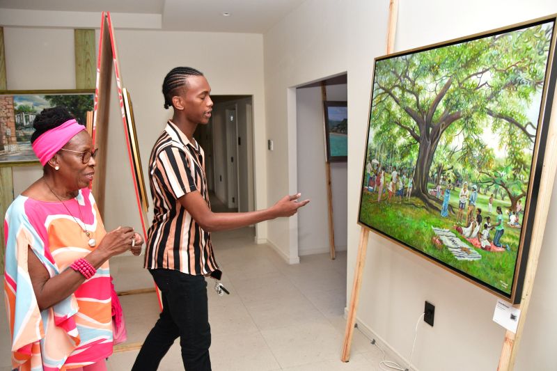 Two people in a bright gallery view a large painting of a park with a big tree.