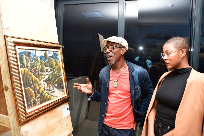 Two people in a gallery discuss a framed landscape painting on a wooden display stand, attentively admiring the art.