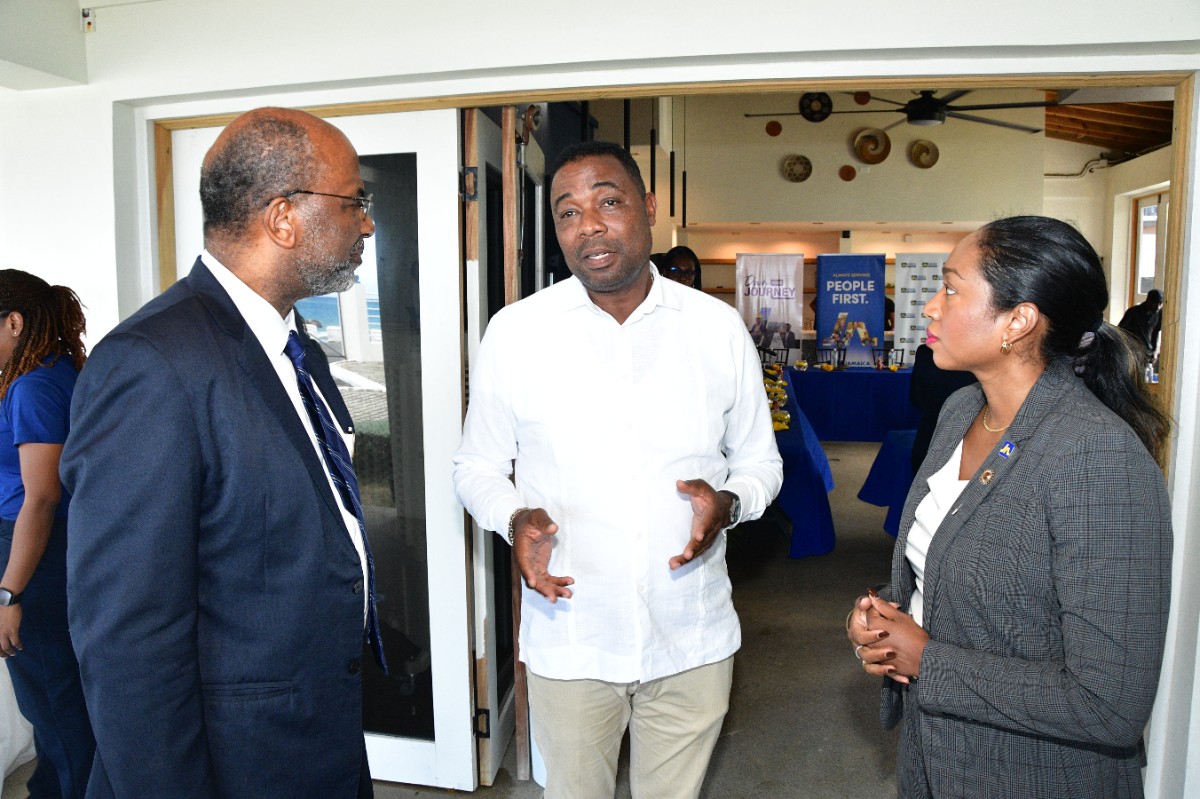 Three adults in a doorway: a man in a dark suit on the left, a man in a white shirt gesturing in the center, and a woman in a gray blazer on the right, conversing at an event.