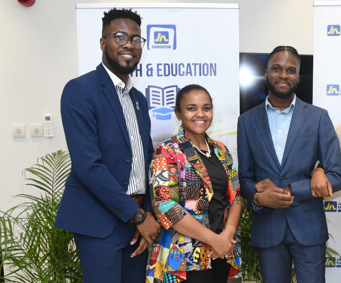 Three smiling professionals pose together in front of a banner at a foundation event, with potted plants nearby.