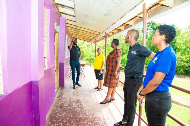DSC09556 - The Jamaica National Group Limited Six adults stand along a covered walkway beside a purple-painted building, with one person gesturing as they discuss renovation or construction plans.