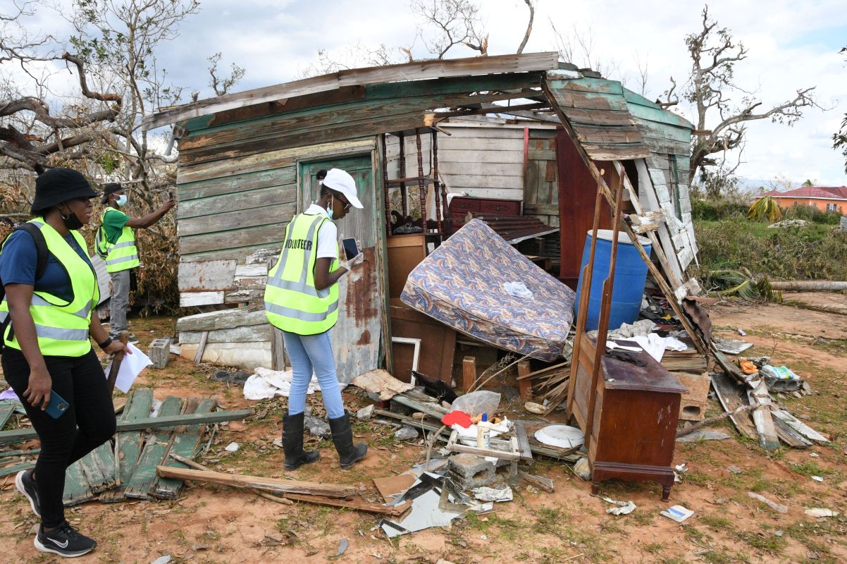 Volunteers in reflective vests survey a dilapidated, debris-strewn shack after a storm disaster scene.
