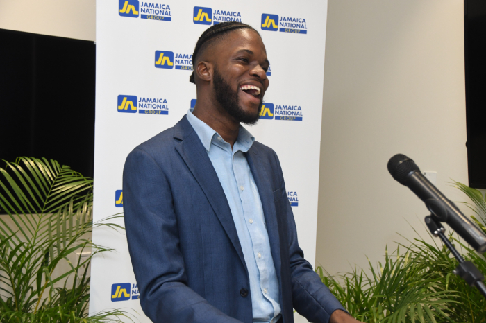 Smiling man in a blue suit speaks at a podium during a Jamaica National Group event, with a microphone nearby and green plants in the foreground.