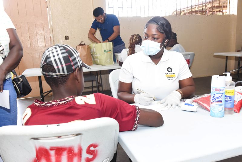 SNP_6745 1 - The Jamaica National Group Limited Nurse in a white uniform with mask and gloves administers an injection to a man at a vaccination table in a clinic setting.