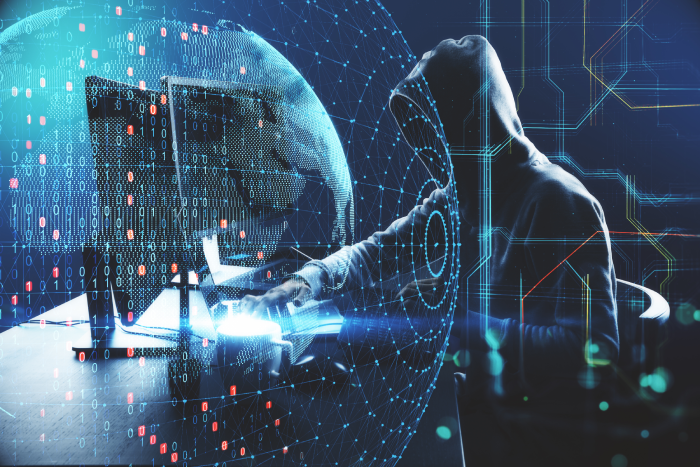 Hooded figure typing at a glowing computer surrounded by a digital globe and data networks, illustrating cybersecurity.